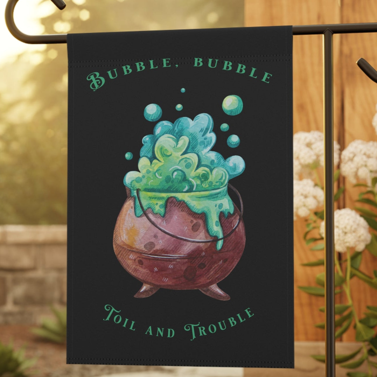 A black garden banner featuring a cartoon illustration of a bubbling cauldron with green liquid, and the text 'Bubble, Bubble, Toil and Trouble' displayed in a garden setting.
