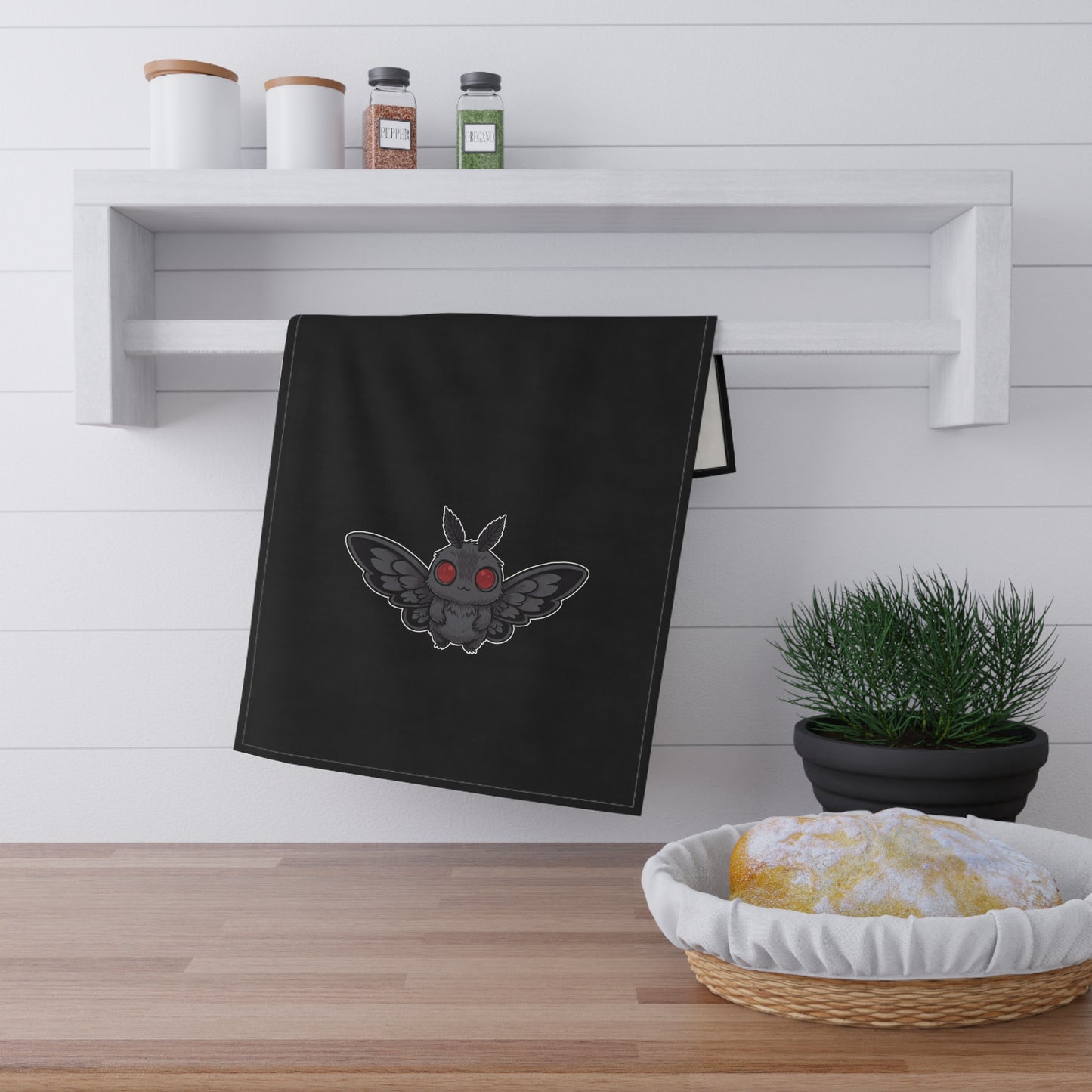 Black towel with a cute chibi Mothman design hanging on a wooden shelf with kitchen items.