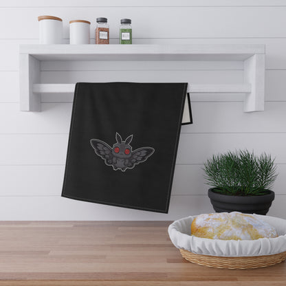 Black towel with a cute chibi Mothman design hanging on a wooden shelf with kitchen items.